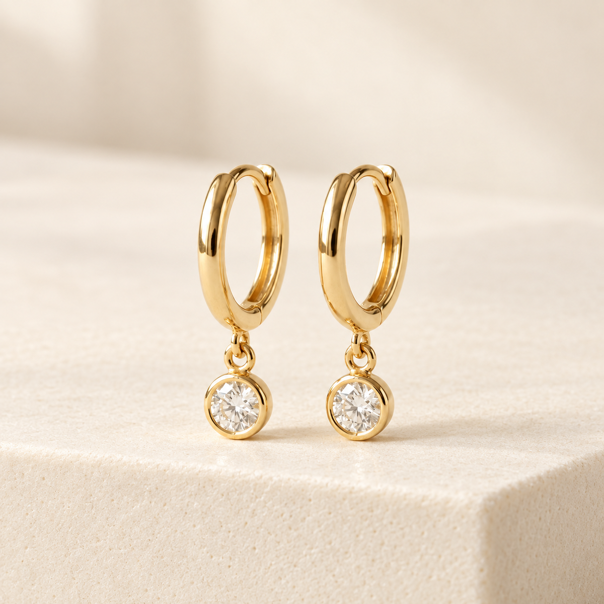 AI-generated visualization — Lab Diamond Bezel Drop Huggies in yellow gold on an ivory studio display.