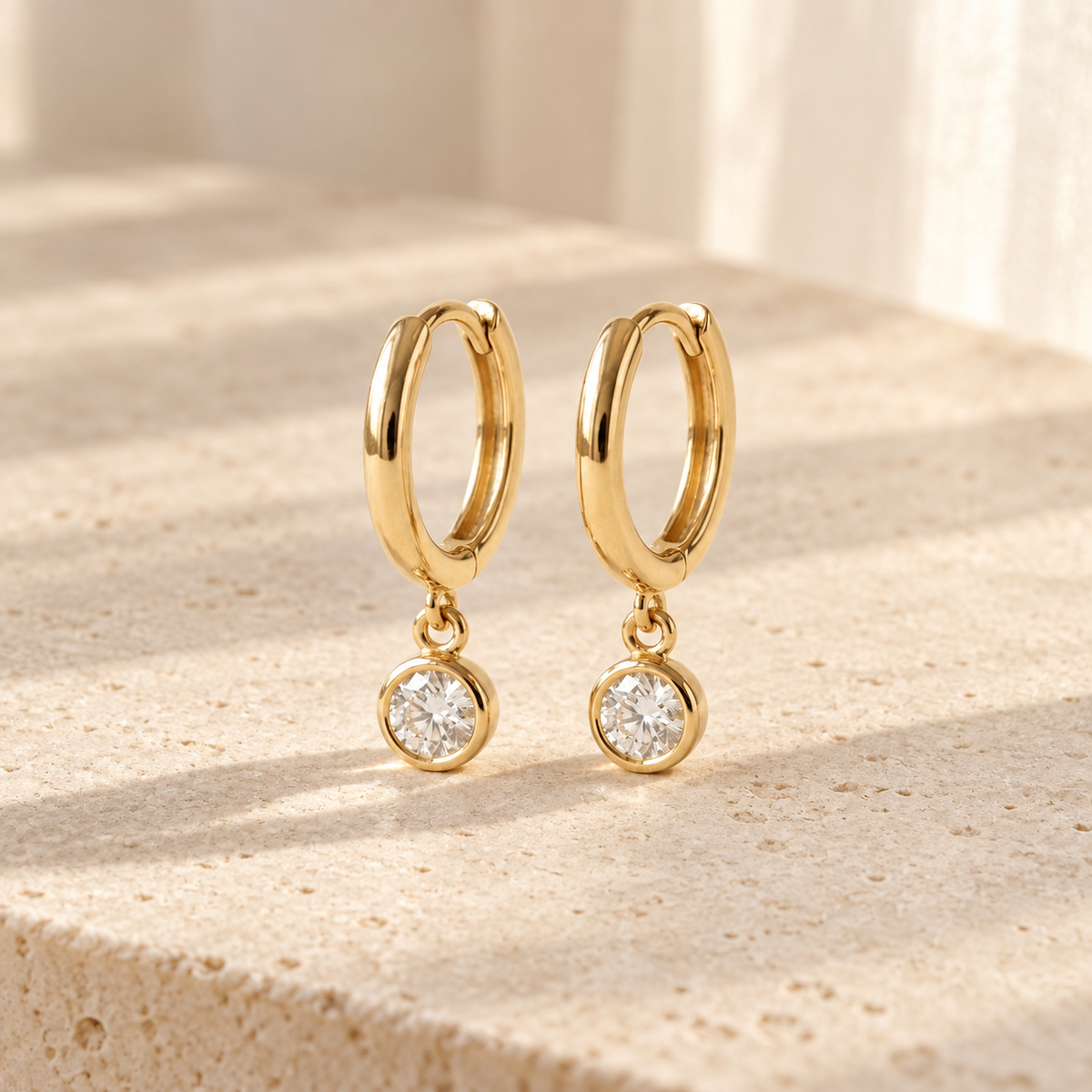 AI-generated visualization — a pair of yellow-gold Bezel Drop Huggies on a warm tabletop in natural window light.
